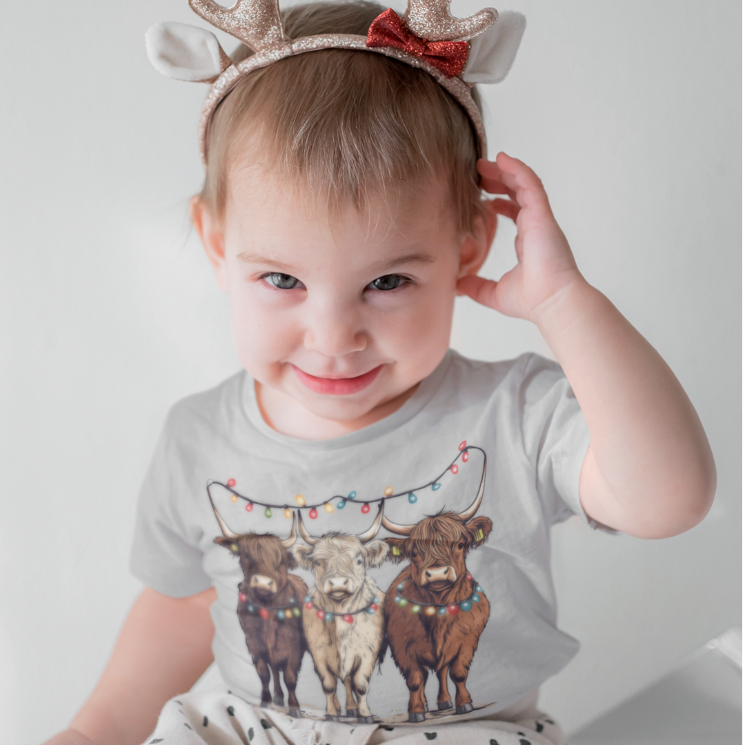Smiling toddler wearing a Christmas T-shirt with three Highland cows decorated in colorful holiday lights.