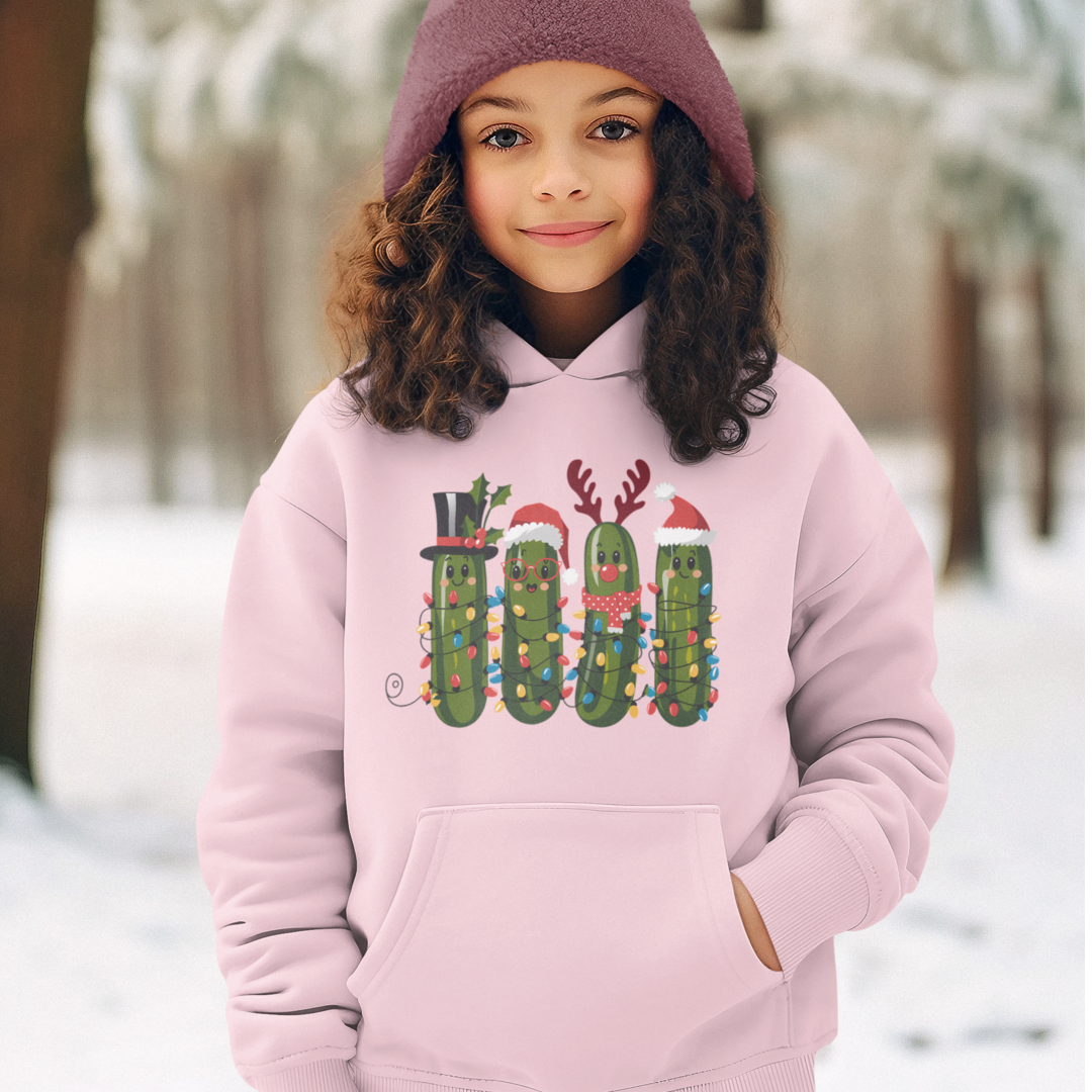 A girl wearing a light pink hoodie with a Christmas pickle graphic, featuring four festive pickles decorated with Santa hats, reindeer antlers, and holiday lights.