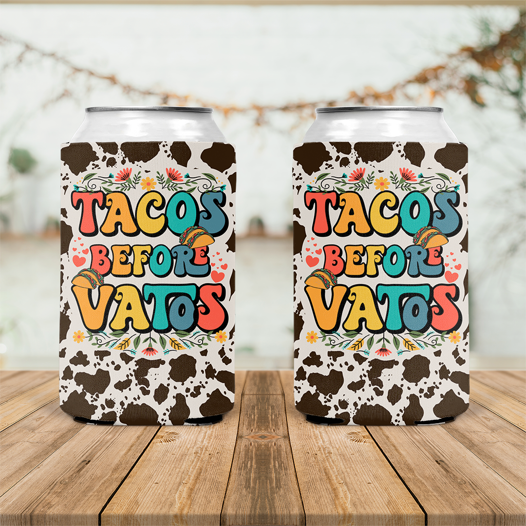 Two cans with cow print sleeves read "TACOS BEFORE VATOS" amidst whimsical taco and flower illustrations on a wooden table, suggesting a fun and festive theme.