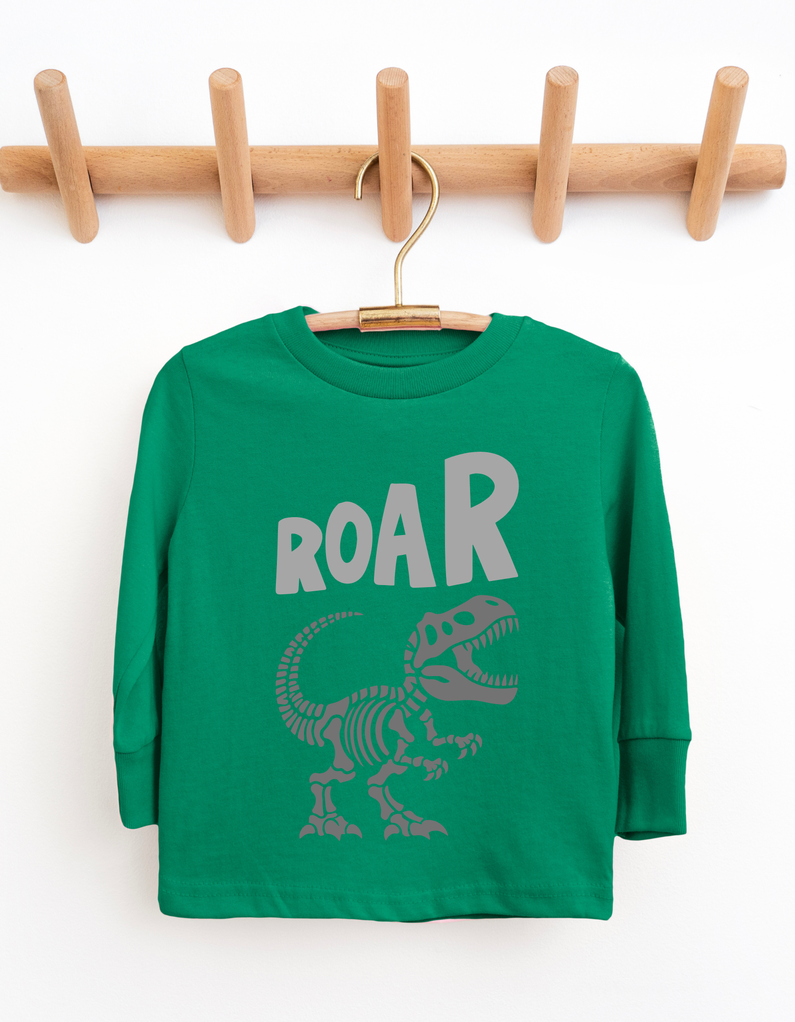 Green children's shirt with dinosaur skeleton and 'ROAR' text on a wooden hanger against a white background