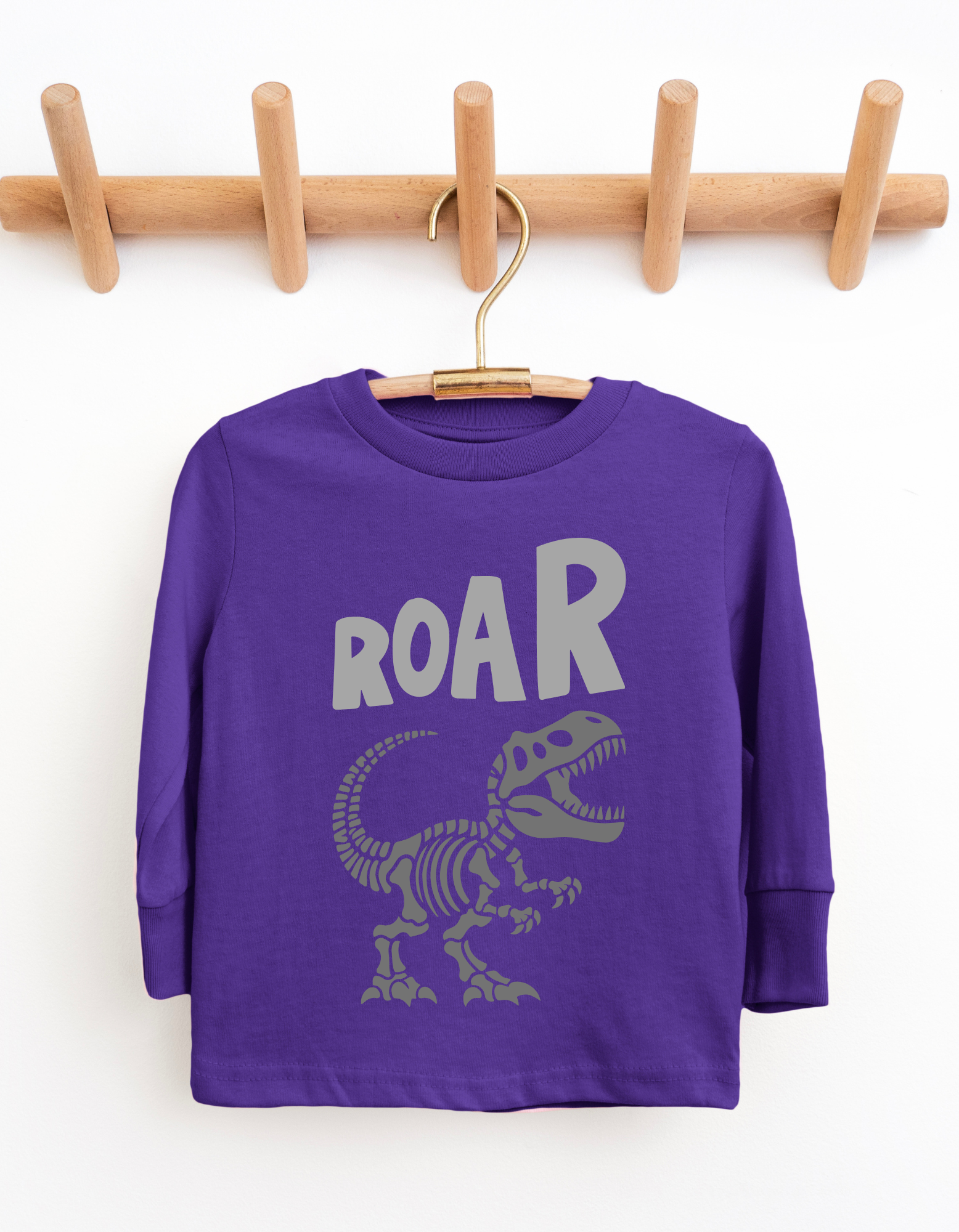 Purple long-sleeve shirt with dinosaur design and 'ROAR' text on a wooden hanger against a white background
