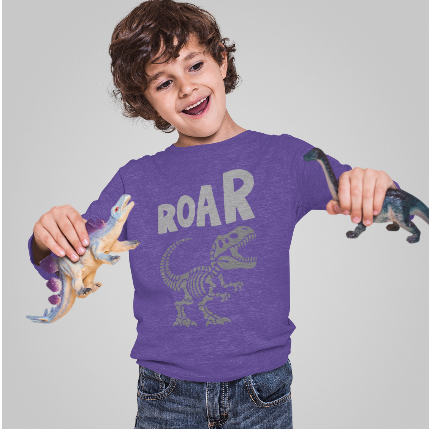 Child wearing a purple shirt with dinosaur design and 'ROAR' text, holding toy dinosaurs against a gray background