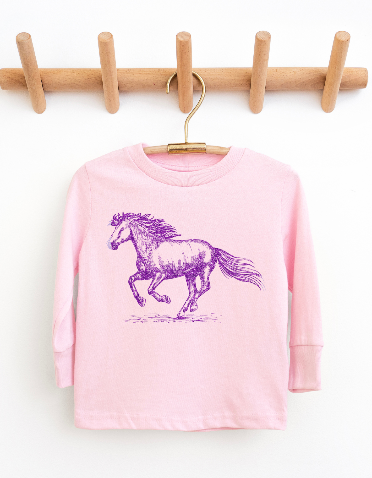 Pink long-sleeve shirt with a purple horse graphic on a wooden hanger against a white background