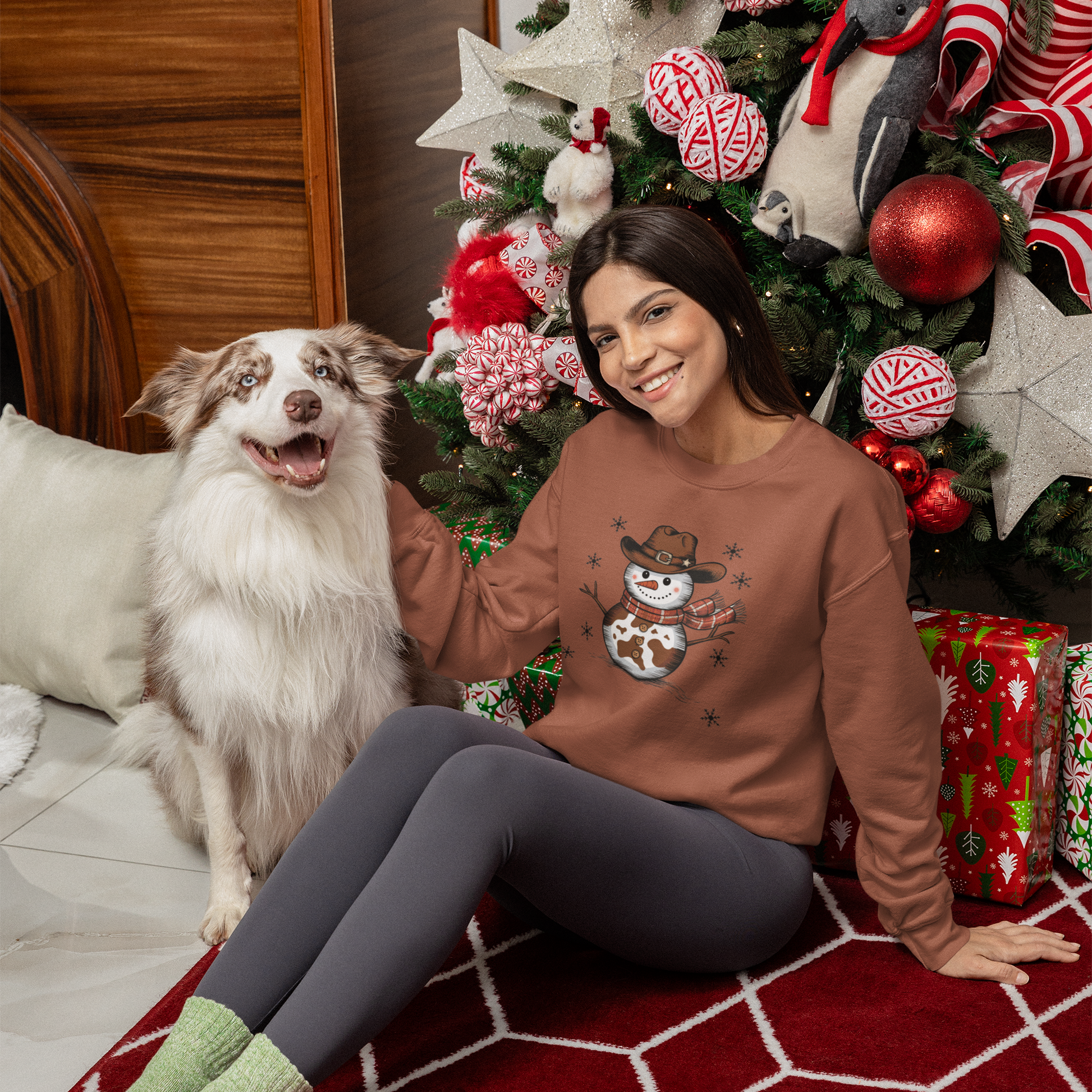 Woman wearing a sweater with a snowman design sitting next to a dog in front of a decorated Christmas tree.