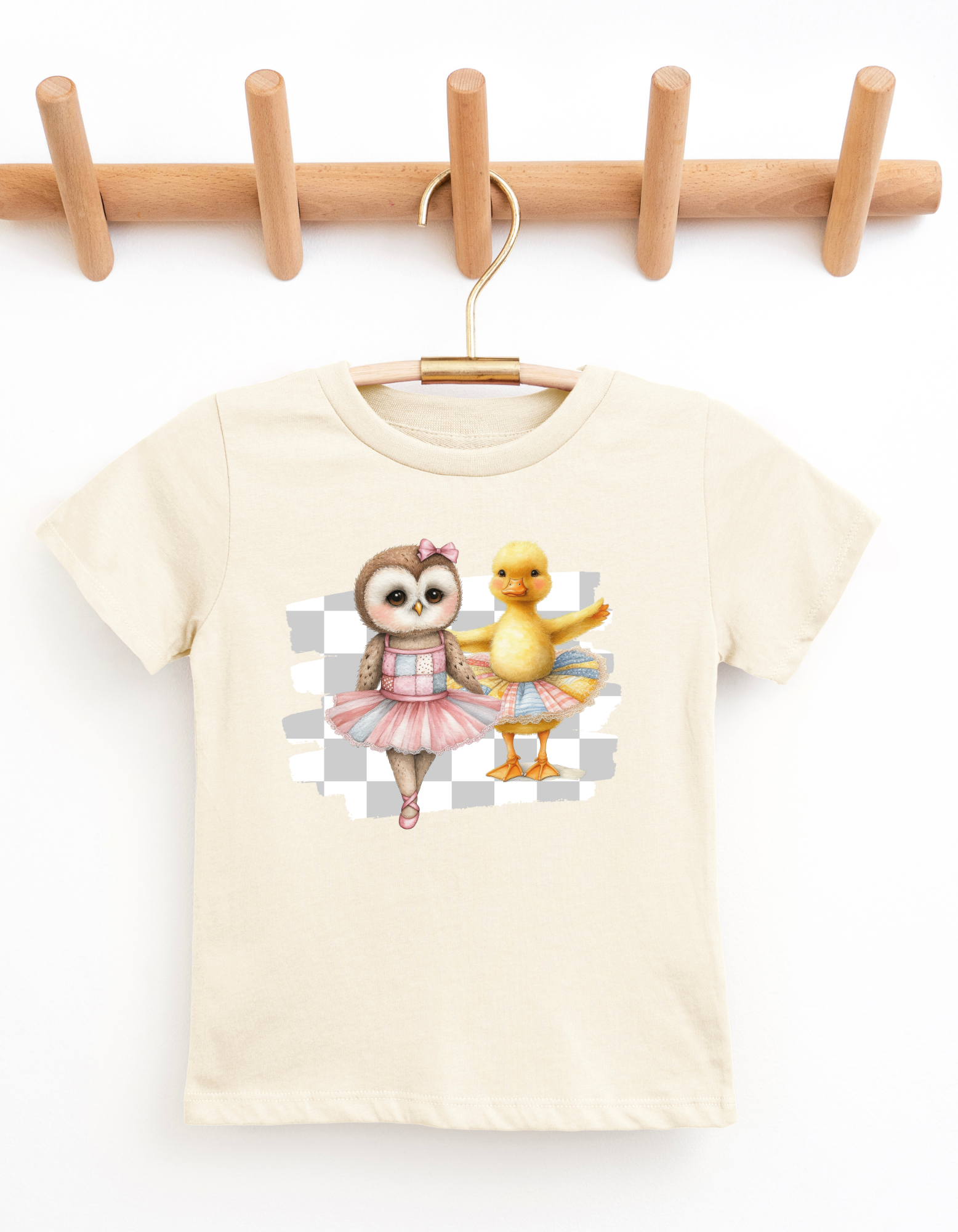 Beige t-shirt with cartoon owl and duck design on a wooden hanger against a white background