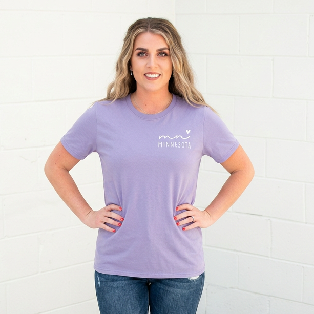 Woman wearing a purple Minnesota t-shirt with text and jeans standing against a white brick wall.