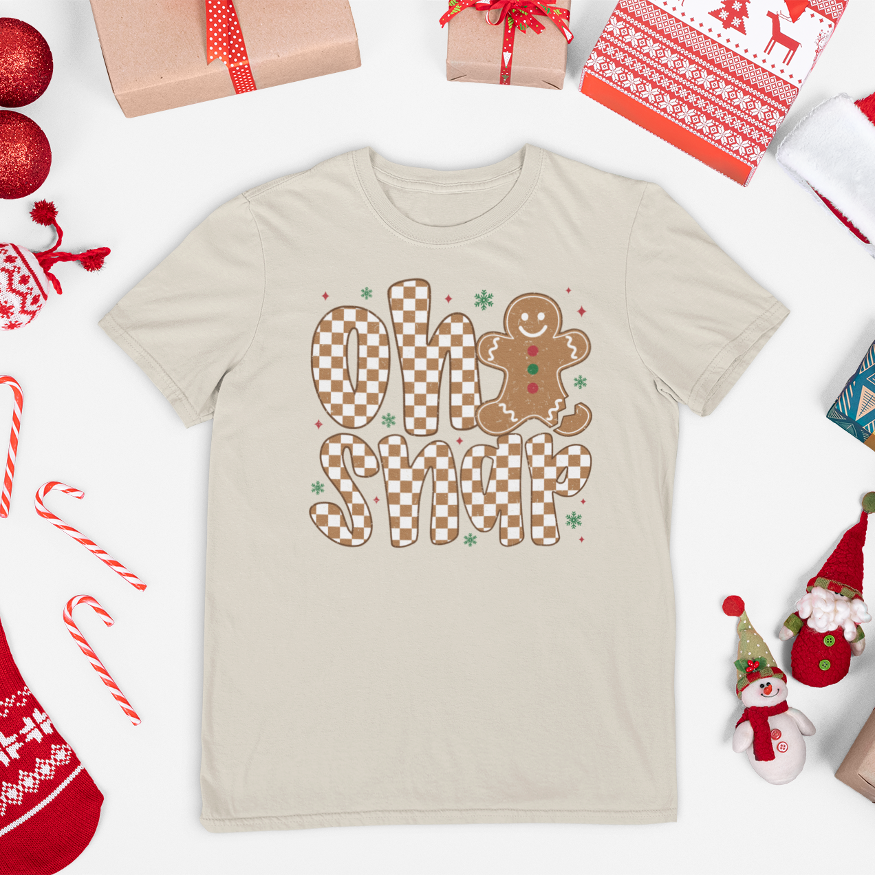 Beige t-shirt with gingerbread design and text, surrounded by Christmas-themed items on a white background