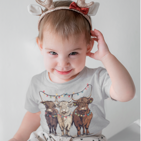 Smiling toddler wearing a Christmas T-shirt with three Highland cows decorated in colorful holiday lights.