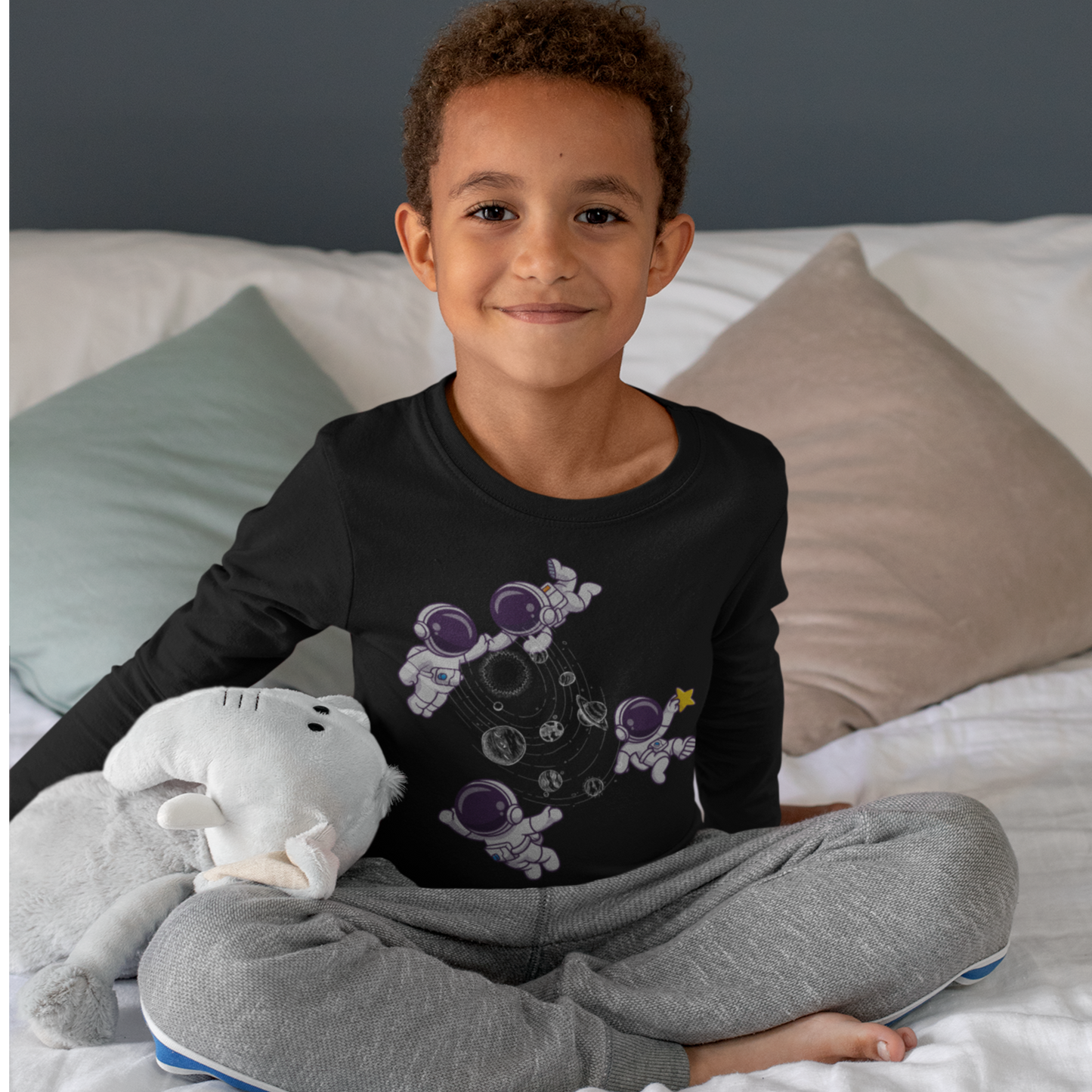 Child wearing a black long-sleeve shirt with a graphic design and gray pants, sitting on a bed.