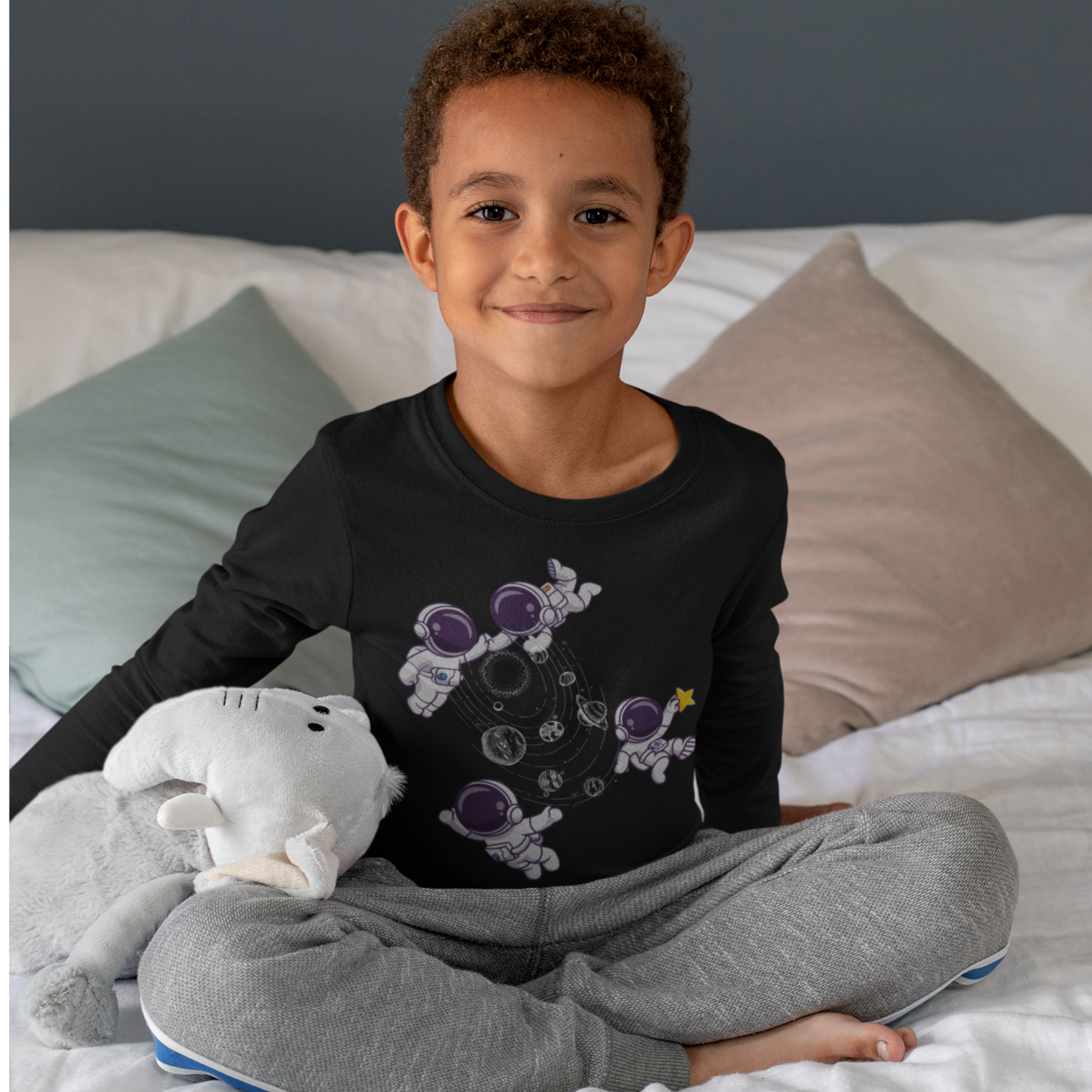 Child wearing a black long-sleeve shirt with a graphic design and gray pants, sitting on a bed.