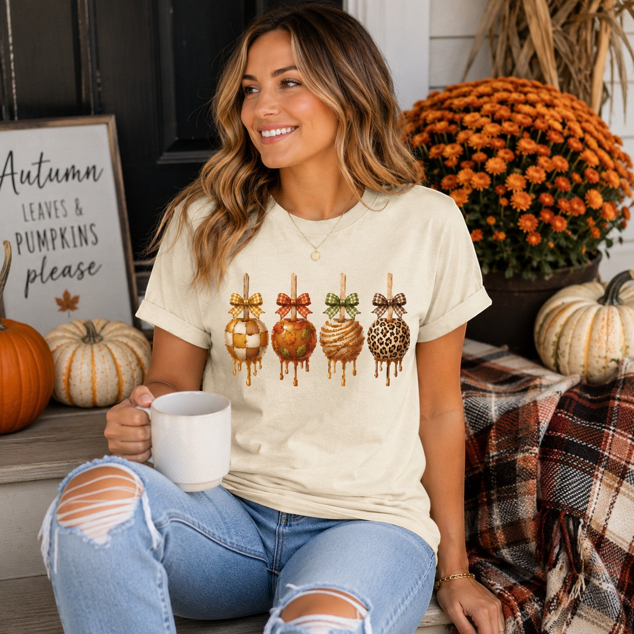 Tea-Shirt Shoppe cream graphic tee featuring four decorated caramel apples with plaid bows, worn by a smiling woman.