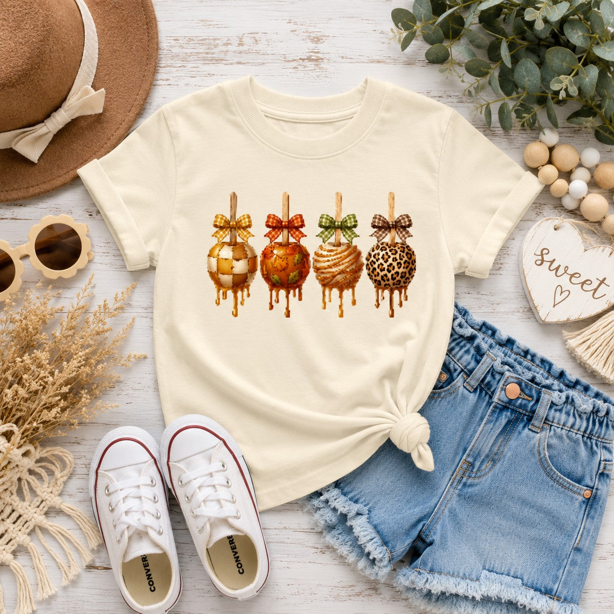 Tea-Shirt Shoppe cream youth graphic tee featuring four caramel apples with bows, styled with denim shorts and hat.