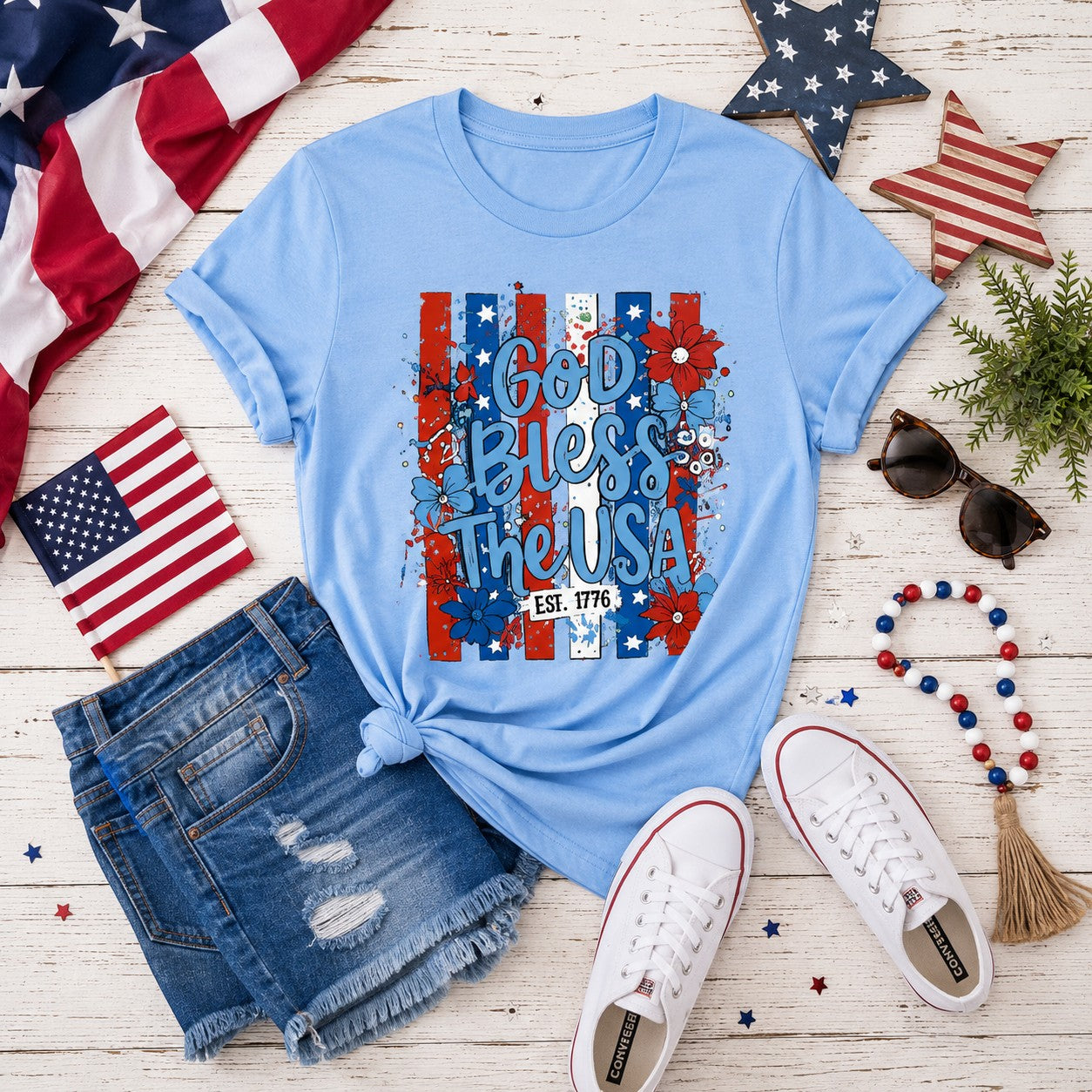 Tea Shirt Shoppe Wholesale light blue graphic tee with "God Bless The USA" text, floral accents, and "Est. 1776" detail.
