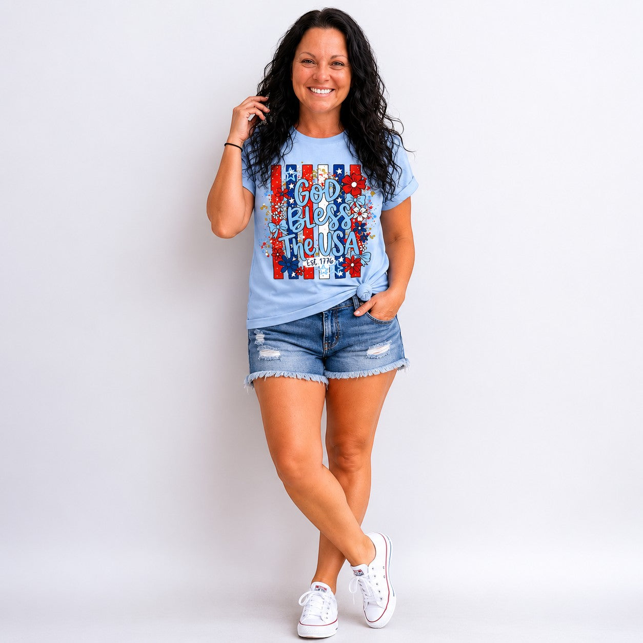 Tea Shirt Shoppe Wholesale light blue graphic tee with "God Bless the USA" text, floral accents, and red, white, and blue stripes. Est. 1776 detail.