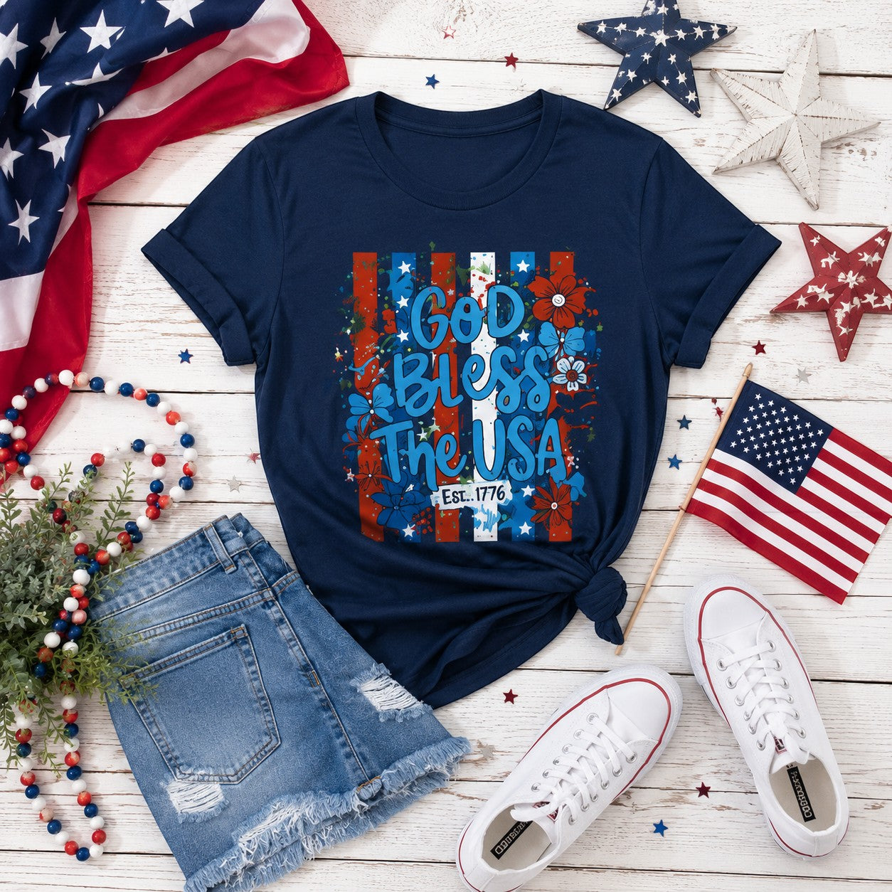 Tea Shirt Shoppe Wholesale navy graphic tee with "God Bless The USA" text, floral accents, and red, white, and blue stripes. Est. 1776 detail.