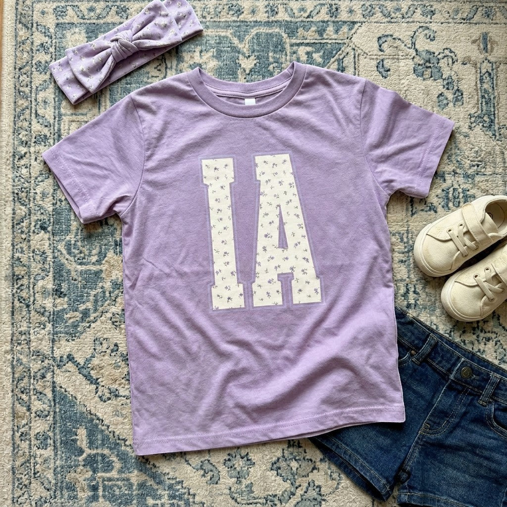 Tea Shirt Shoppe Wholesale lavender cotton t-shirt with "IA" floral graphic, paired with denim shorts and sneakers.