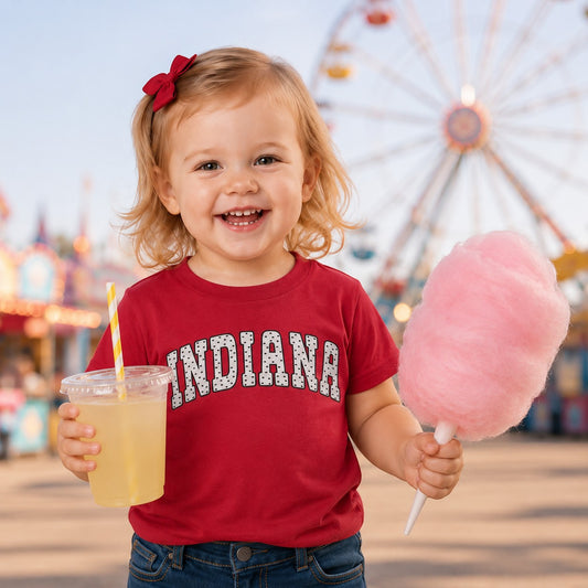 Toddler wearing a maroon Tea Shirt Shoppe Wholesale Indiana graphic tee with polka dot text at a carnival