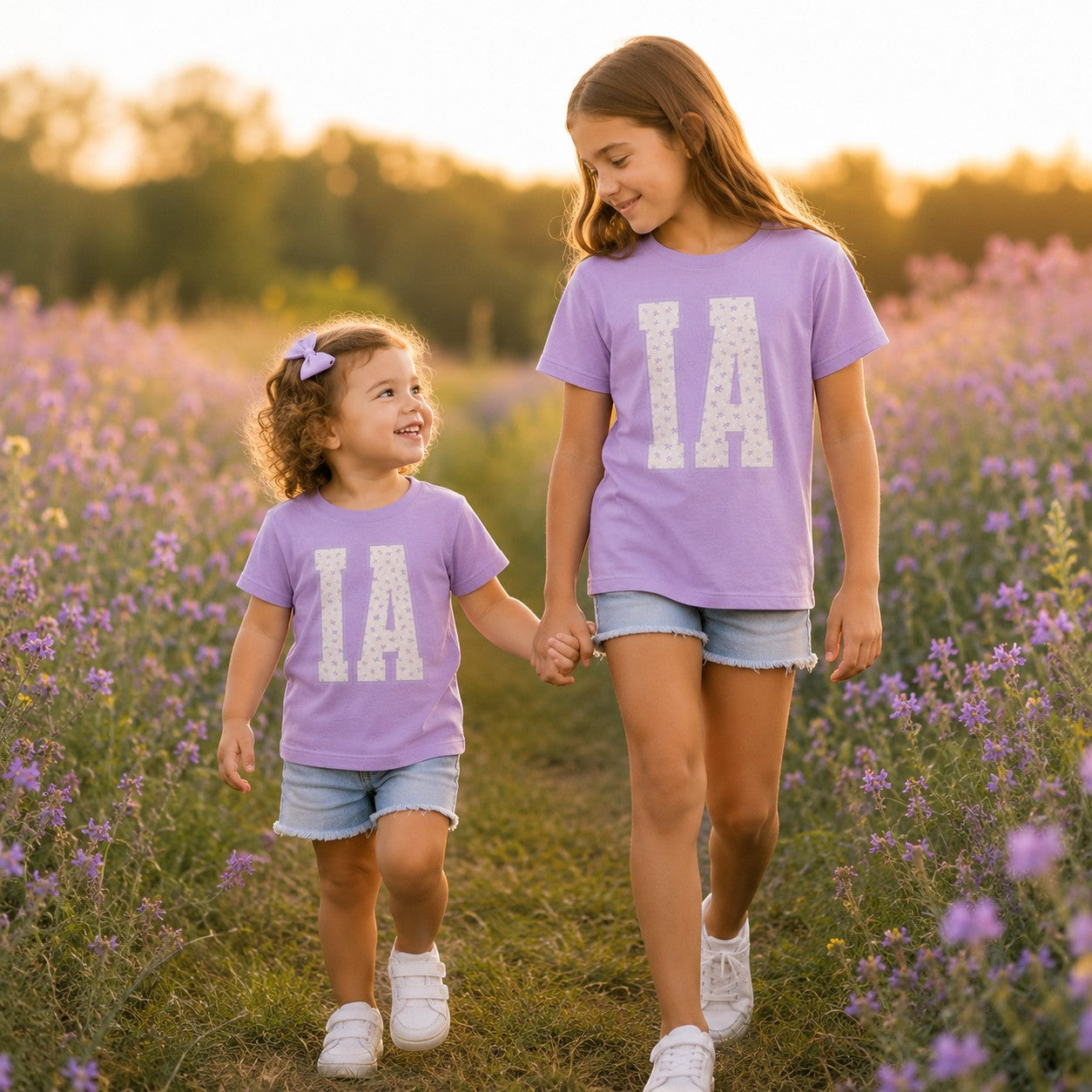 Two girls wearing Tea Shirt Shoppe Wholesale lavender cotton t-shirts with floral IA varsity graphic in a flower field