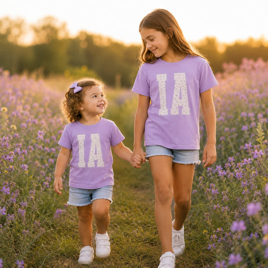 Two girls wearing Tea Shirt Shoppe Wholesale lavender cotton t-shirts with floral IA varsity graphic in a flower field