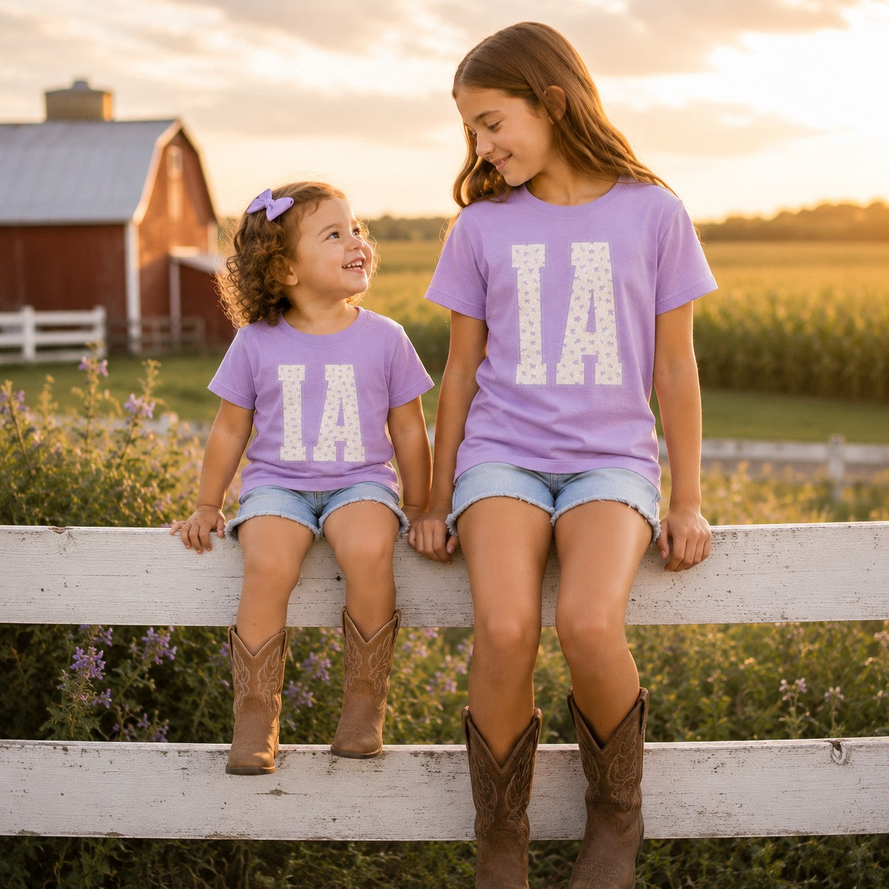 Two girls wearing Tea Shirt Shoppe Wholesale lavender cotton t-shirts with floral IA varsity graphic on a farm fence.