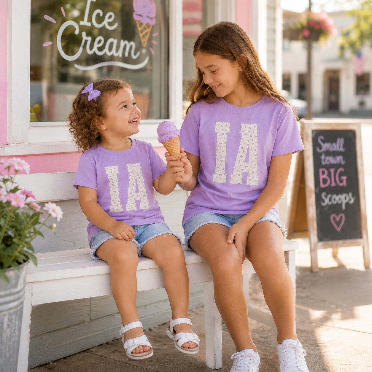 Two girls wearing Tea Shirt Shoppe Wholesale lavender cotton t-shirts with floral IA varsity graphic on a bench.