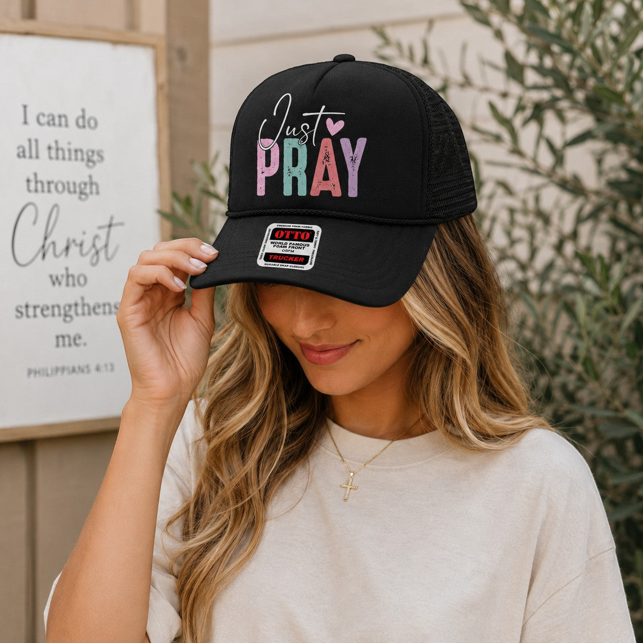 Tea Shirt Shoppe Wholesale black trucker hat with Just Pray graphic in pastel colors, worn by a woman outdoors.