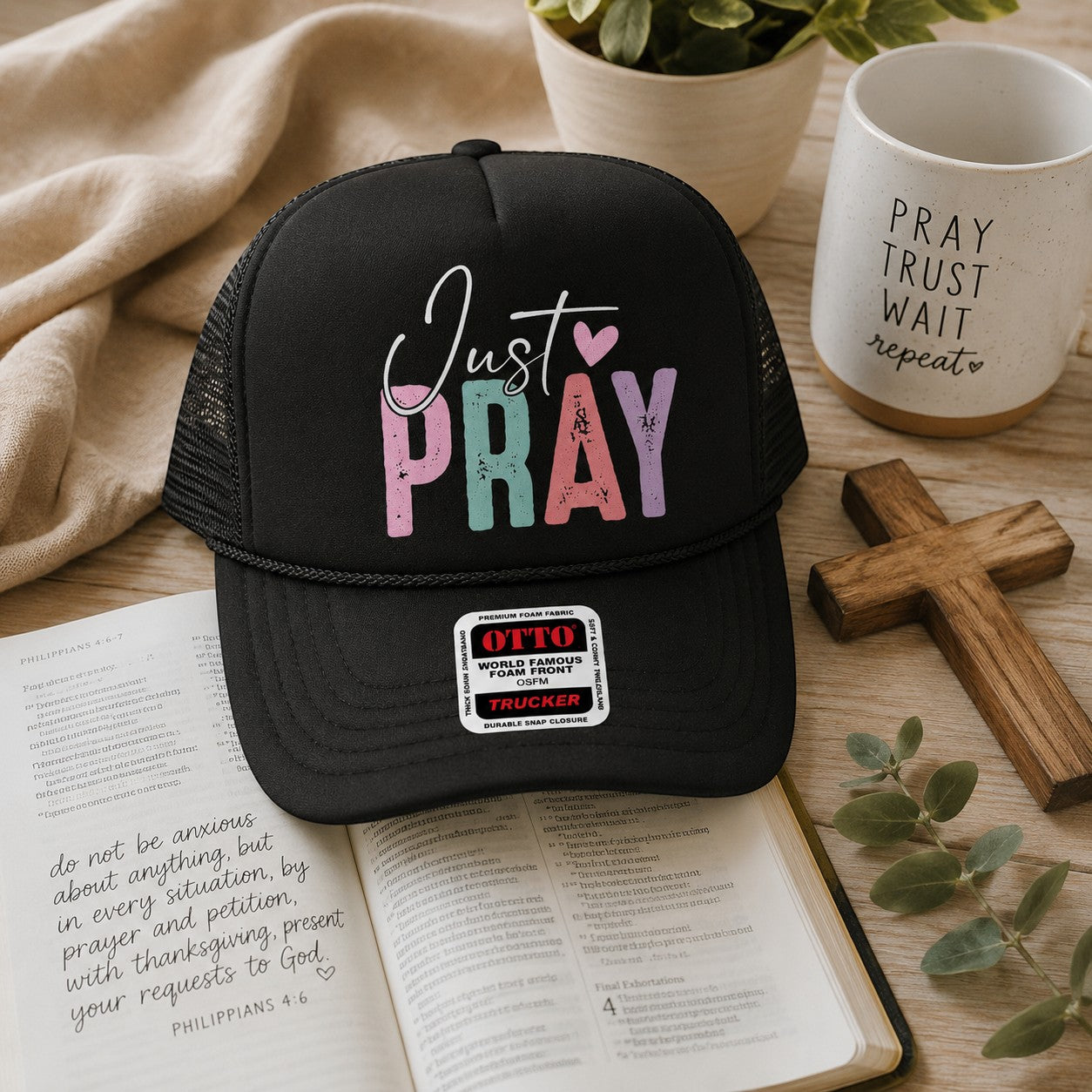 Tea Shirt Shoppe Wholesale black trucker hat with Just Pray graphic in pastel distressed lettering on a bible desk.