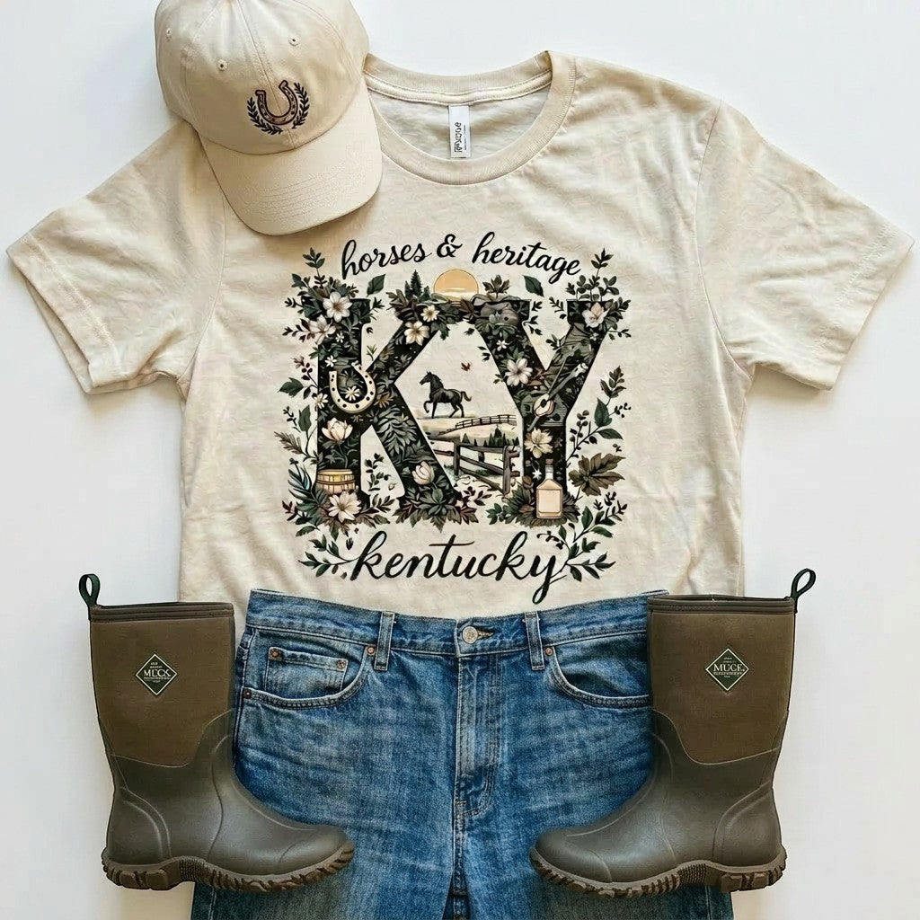 Tea Shirt Shoppe Wholesale cream graphic tee with "KY" design featuring horses, fences, bourbon barrels, and floral elements. Text reads "horses & heritage kentucky".