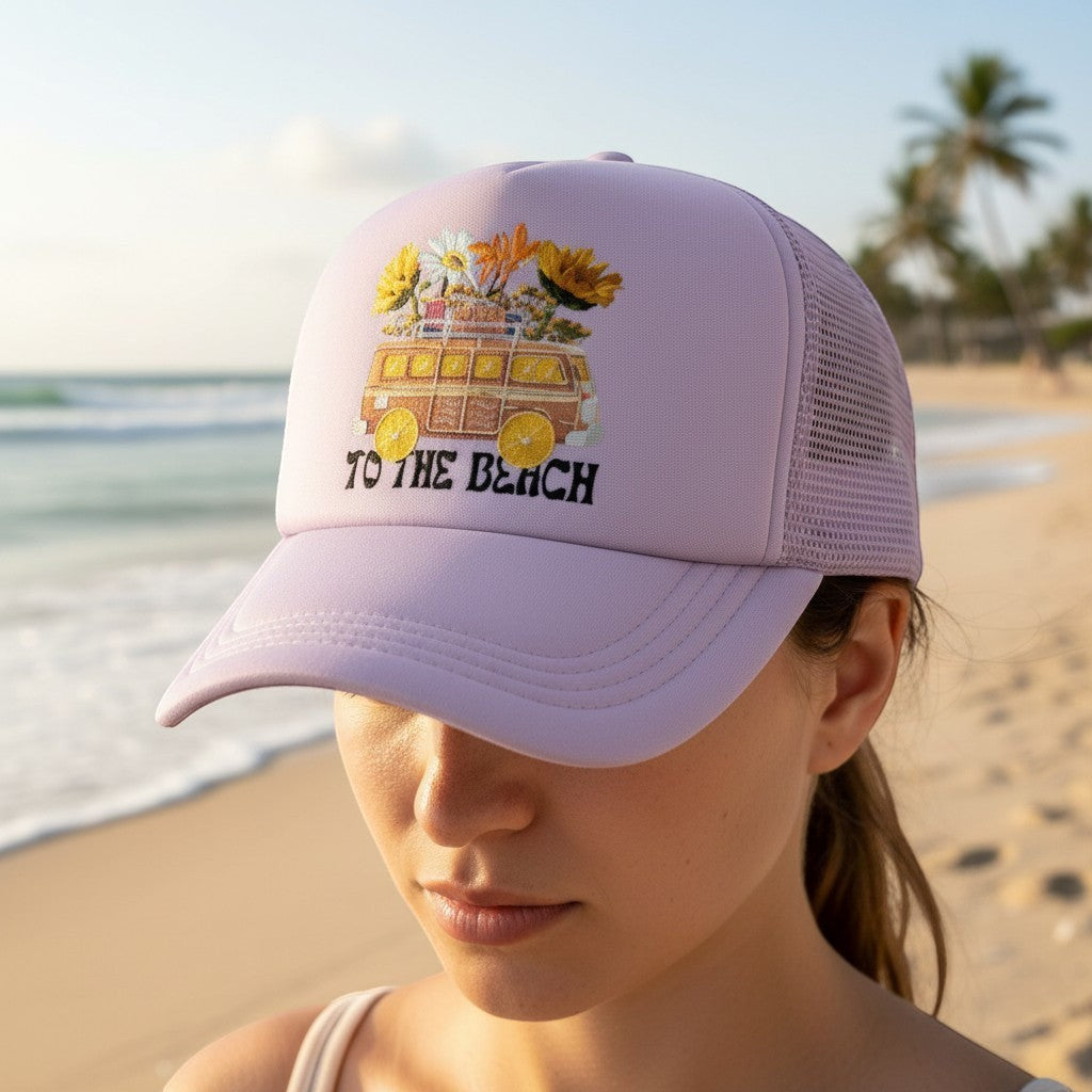 Tea Shirt Shoppe Wholesale lavender trucker hat with embroidered van and flowers graphic and "To The Beach" text.