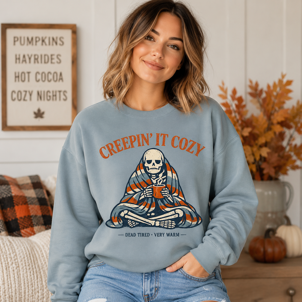 Woman wearing a light slate Tea Shirt Shoppe Wholesale graphic sweatshirt featuring a skeleton wrapped in a blanket.