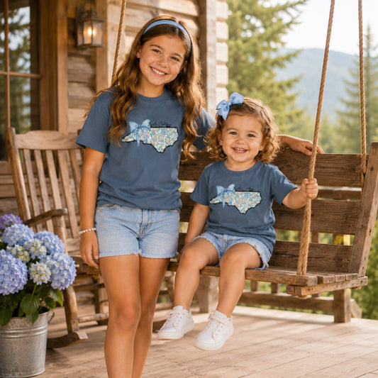 Two girls wearing Tea Shirt Shoppe Wholesale indigo cotton t-shirts with a floral North Carolina state graphic and bow.