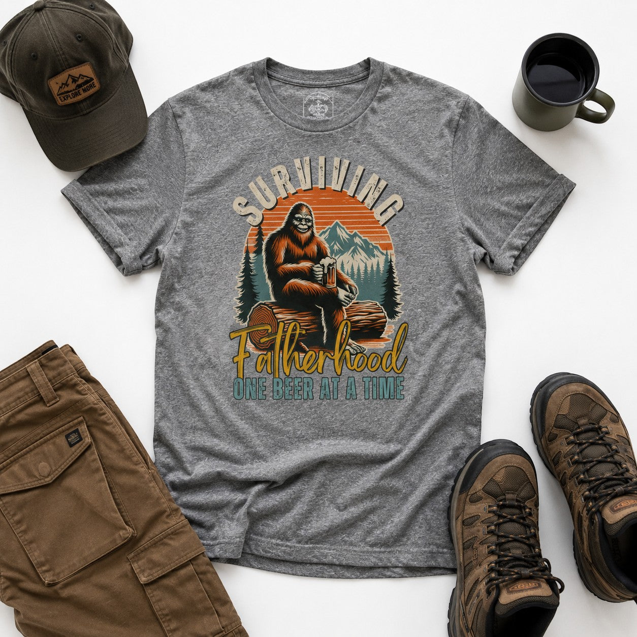 Tea Shirt Shoppe Wholesale grey graphic tee with Bigfoot sitting on a log, holding a beer, with mountains and sunset graphic. Text reads "Surviving Fatherhood One Beer At A Time."