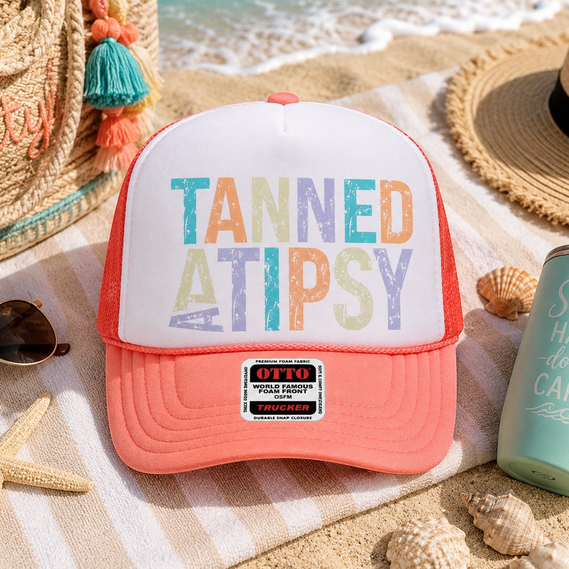 Tea Shirt Shoppe Wholesale Tanned And Tipsy trucker hat with coral foam front and mesh back on a beach towel.
