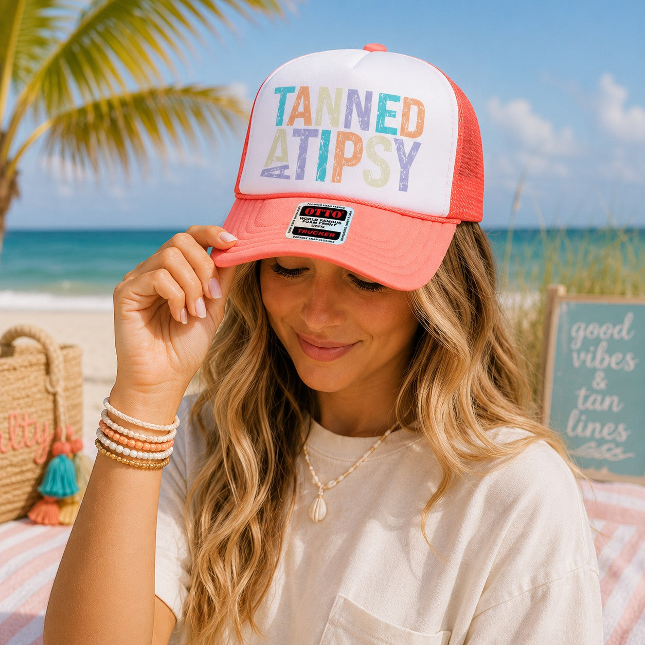 Tea Shirt Shoppe Wholesale Tanned And Tipsy trucker hat with coral mesh back and white foam front on a beach model.