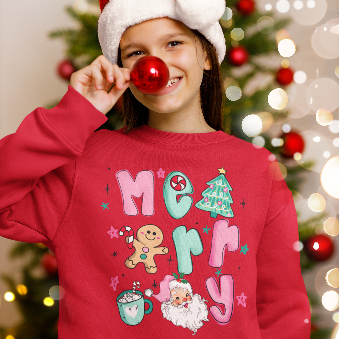 Child wearing a red Christmas-themed sweatshirt with decorations, holding a red ornament in front of a blurred Christmas tree.